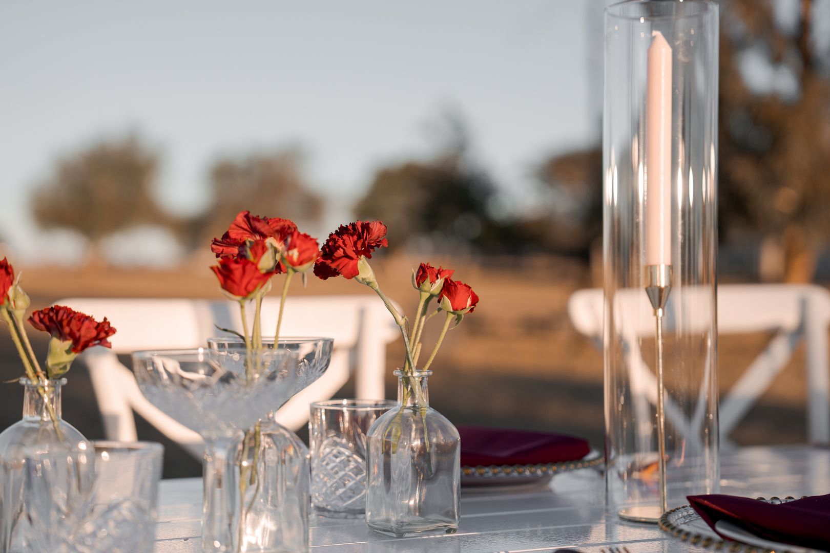 Small glass vases with red flowers on a sunlit outdoor dining table, with white candles and blurred background