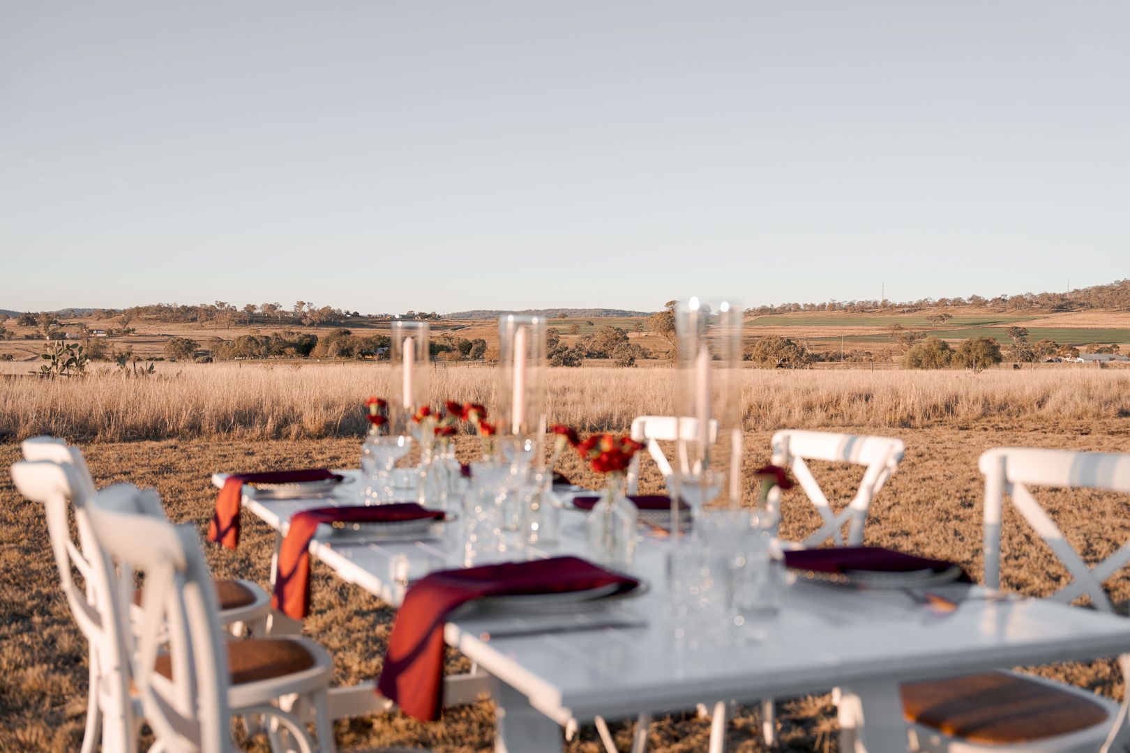 Outdoor dining table set with white chairs in a dry field, glasses and red napkins.