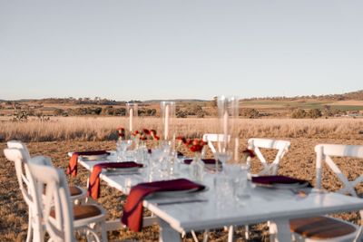 Outdoor dining table set with white chairs in a dry field, glasses and red napkins.
