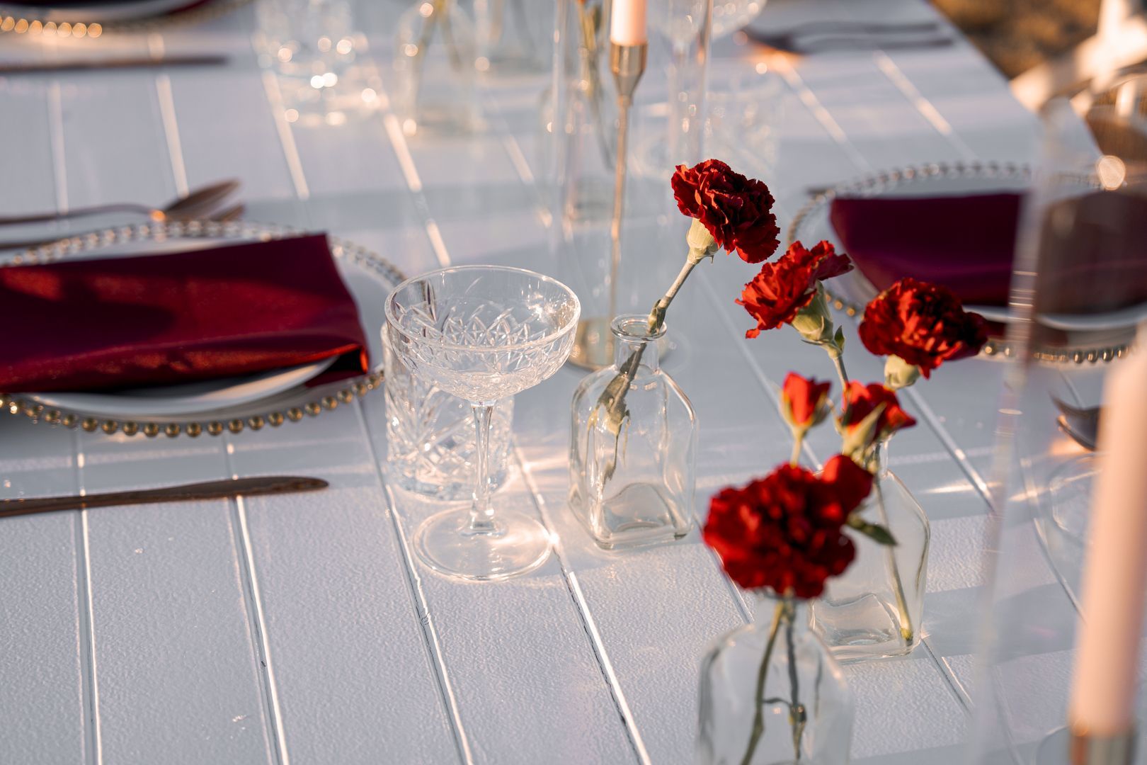 Elegant table setting with red napkins, clear glassware, and small red floral centerpieces.