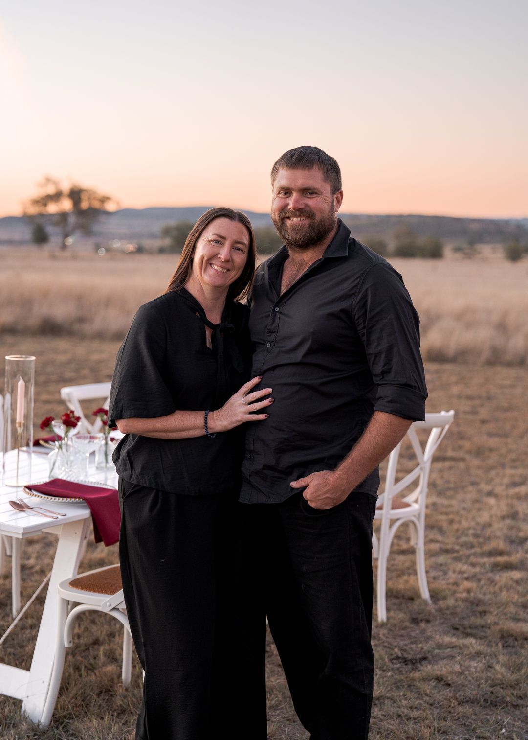 Couple posing outdoors beside a set dining table at sunset, smiling and embracing