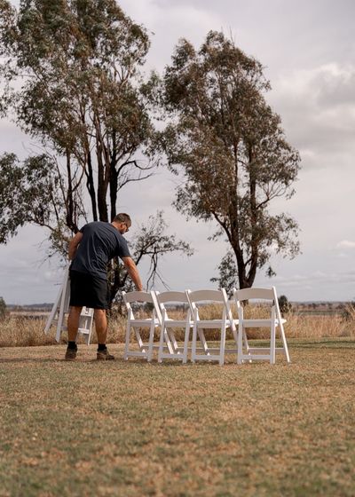 Person setting up white folding chairs outdoors in a grassy field with trees in the background