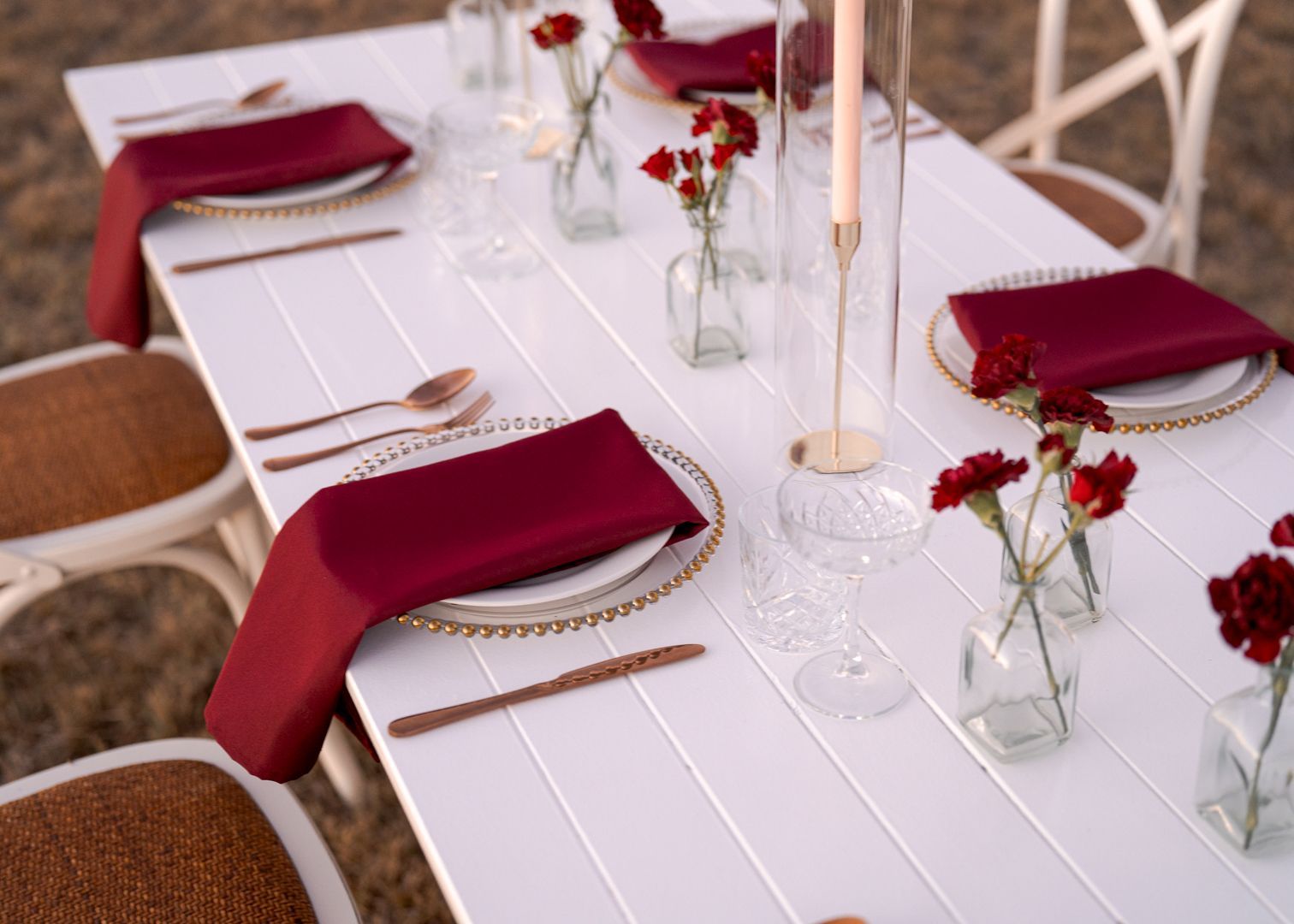 Elegant outdoor table setting with burgundy napkins, gold flatware, and white candles.