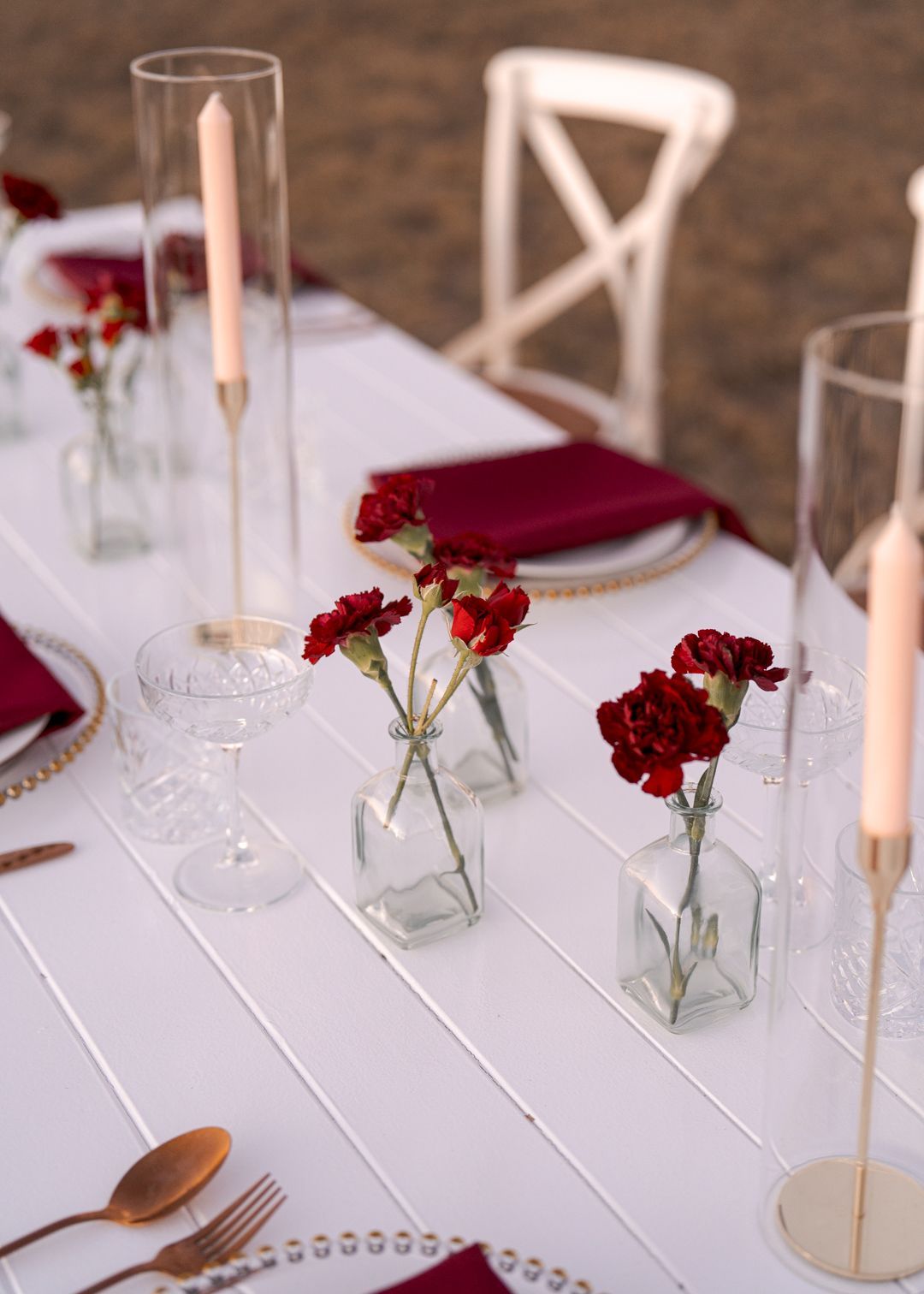Elegant white table setting with red flowers in glass vases and burgundy napkins, lit by tall candles