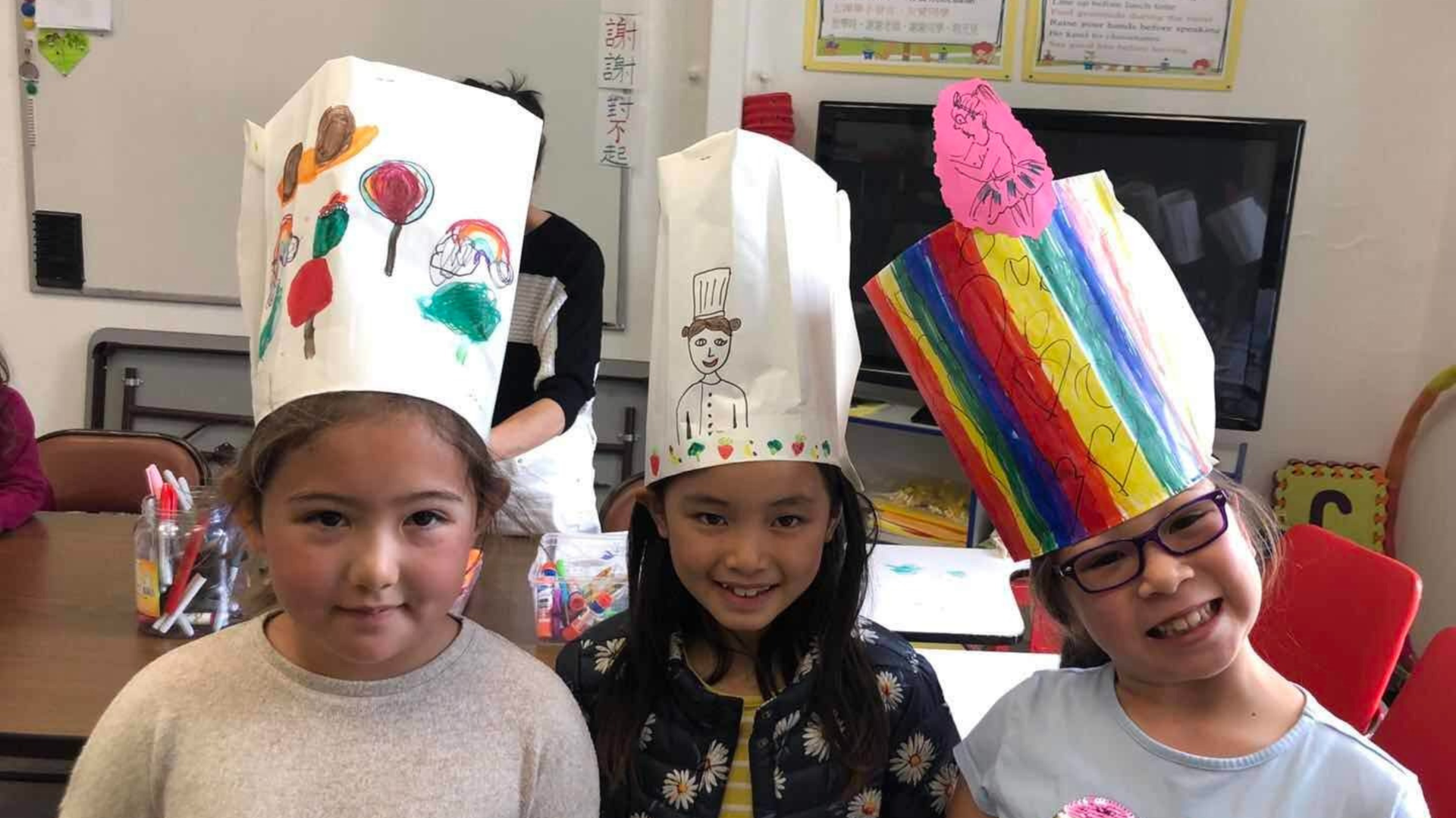Three children wearing tall decorated paper hats sit in a classroom, smiling at the camera.