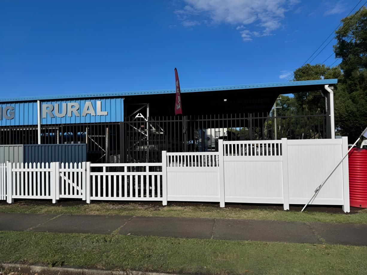 A white picket fence in front of a blue rural building with a black gate.