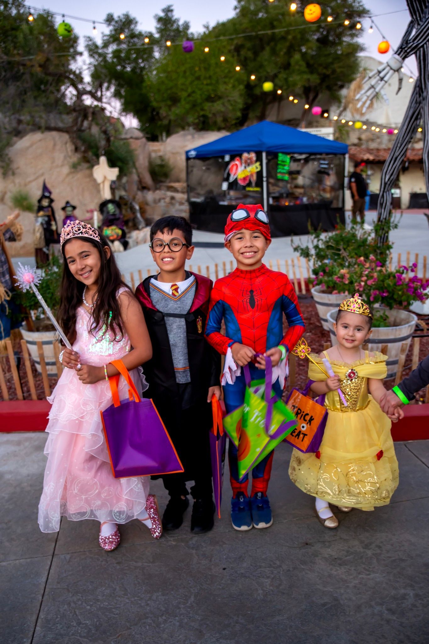 Four children in colorful costumes holding treat bags at an outdoor Halloween event