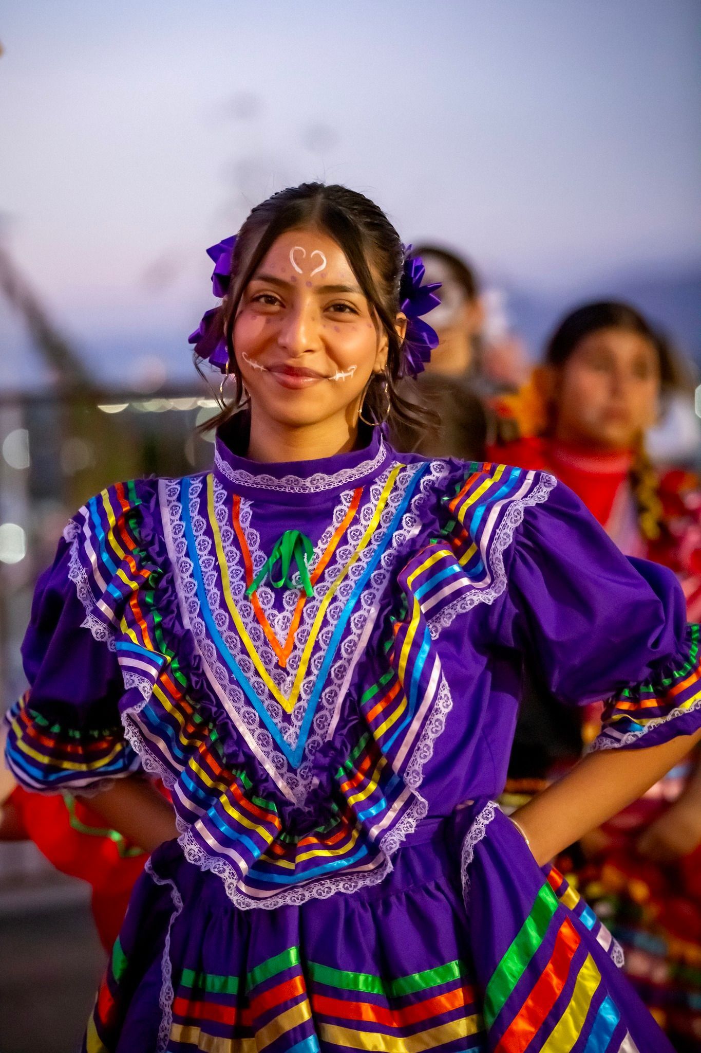Woman in a bright purple embroidered dress standing outdoors at dusk, smiling with hands on hips.