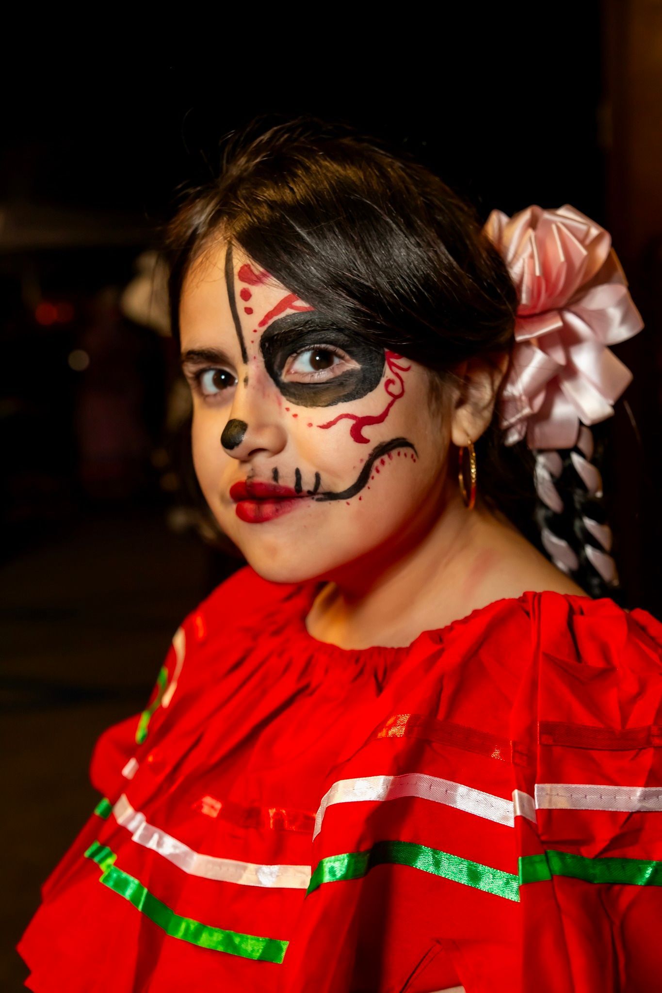 Child in red dress with Día de los Muertos face paint and braided hair with pink flowers