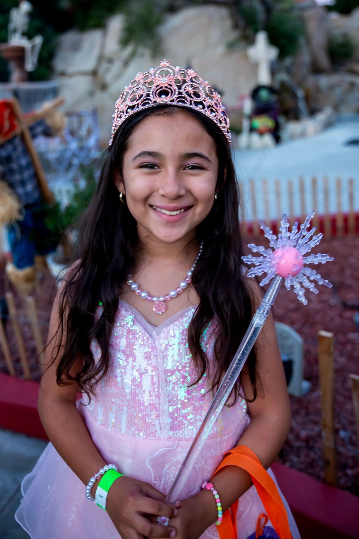 Smiling child in a pink dress and tiara holding a wand and colorful bag at a festive event