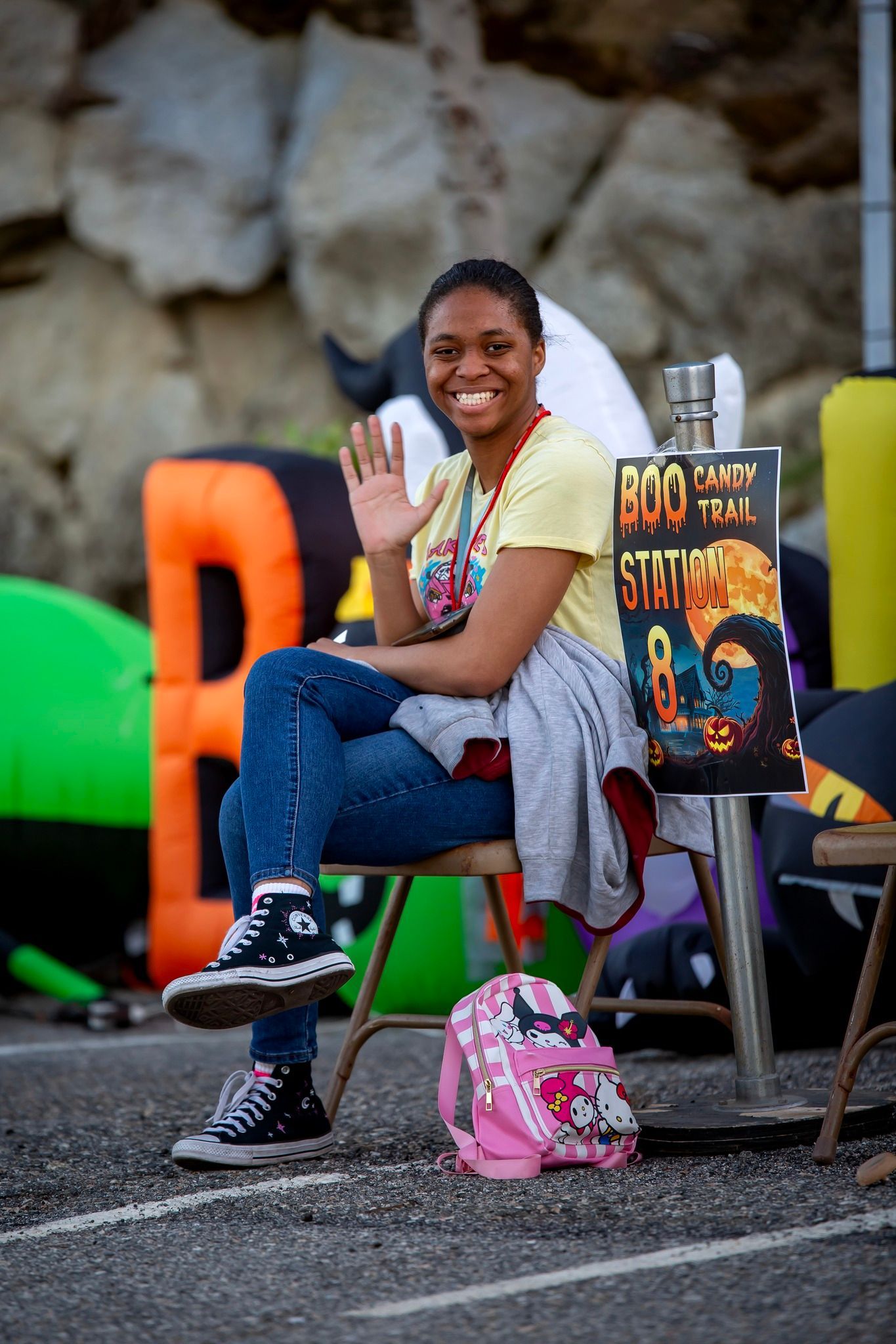 Smiling child seated outdoors on a chair, holding snacks beside a pink backpack and colorful artwork display