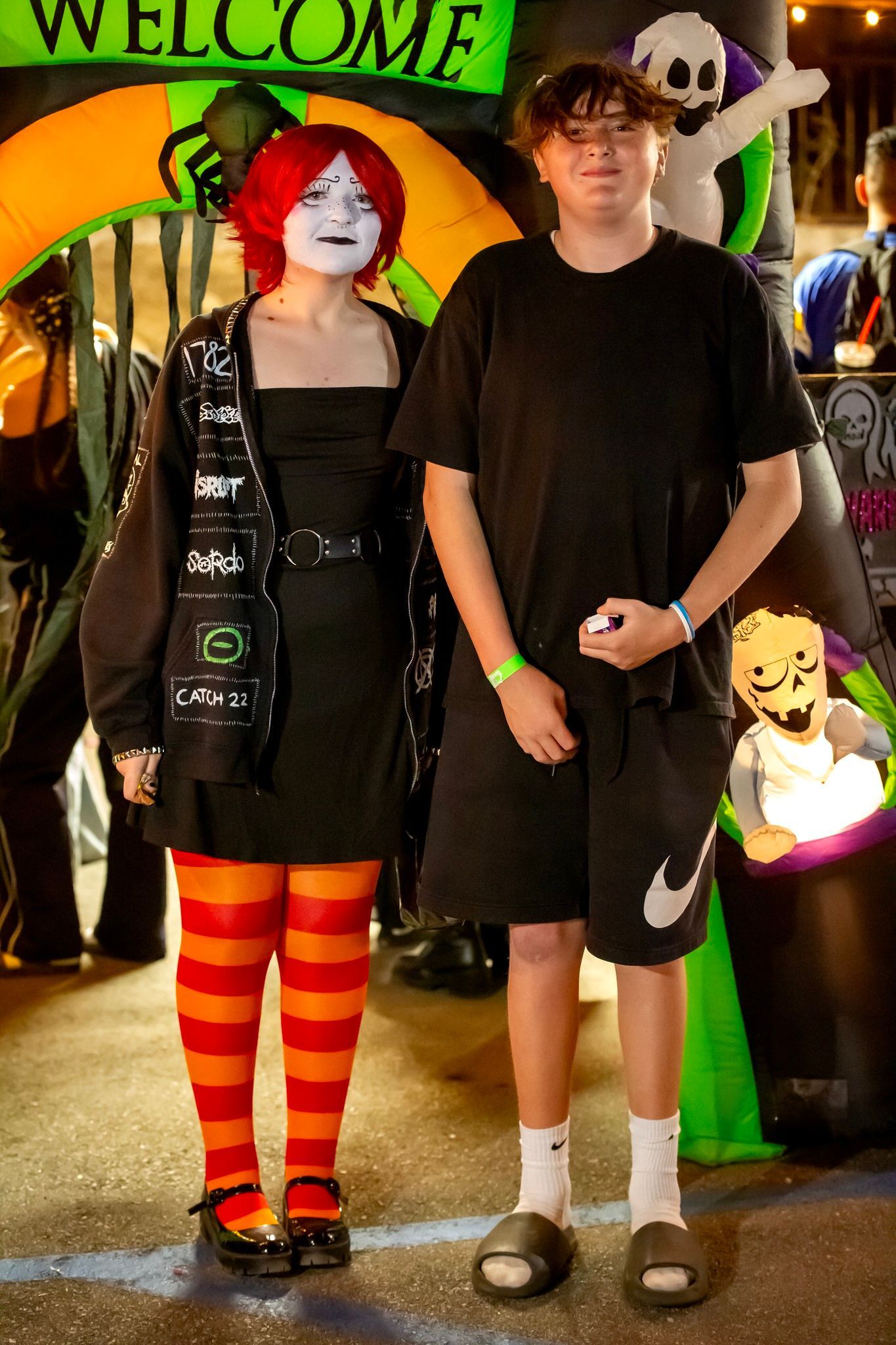 Two costumed people posing at a Halloween display with a green “Welcome” sign and pumpkins.