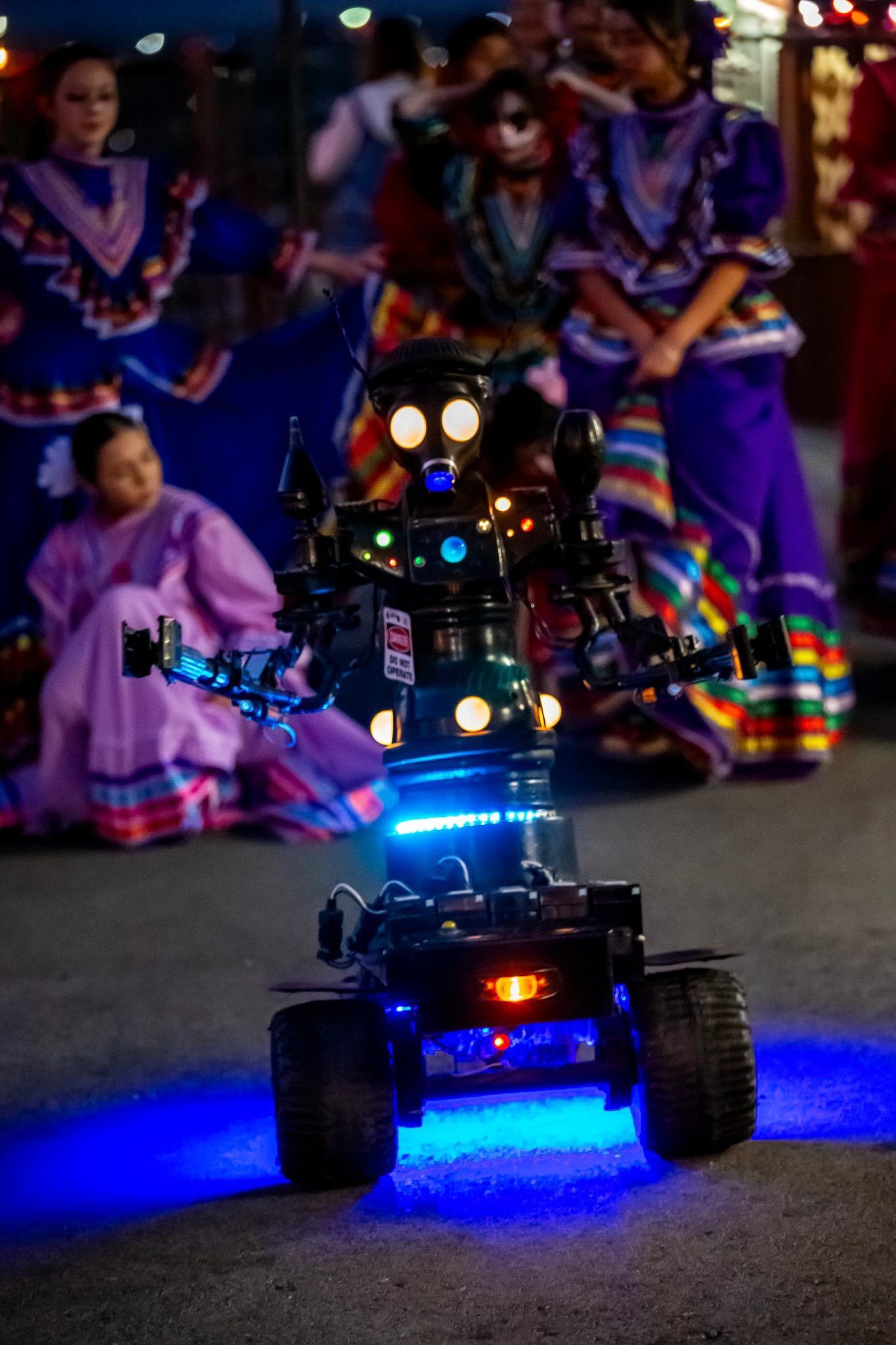 Small blue-lit toy car on a street at night with blurred dancers in colorful costumes behind it