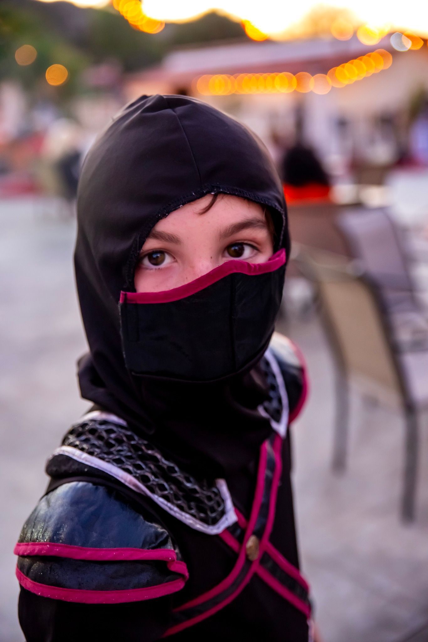 Person in black and pink hood and face covering, standing outdoors with blurred lights in the background