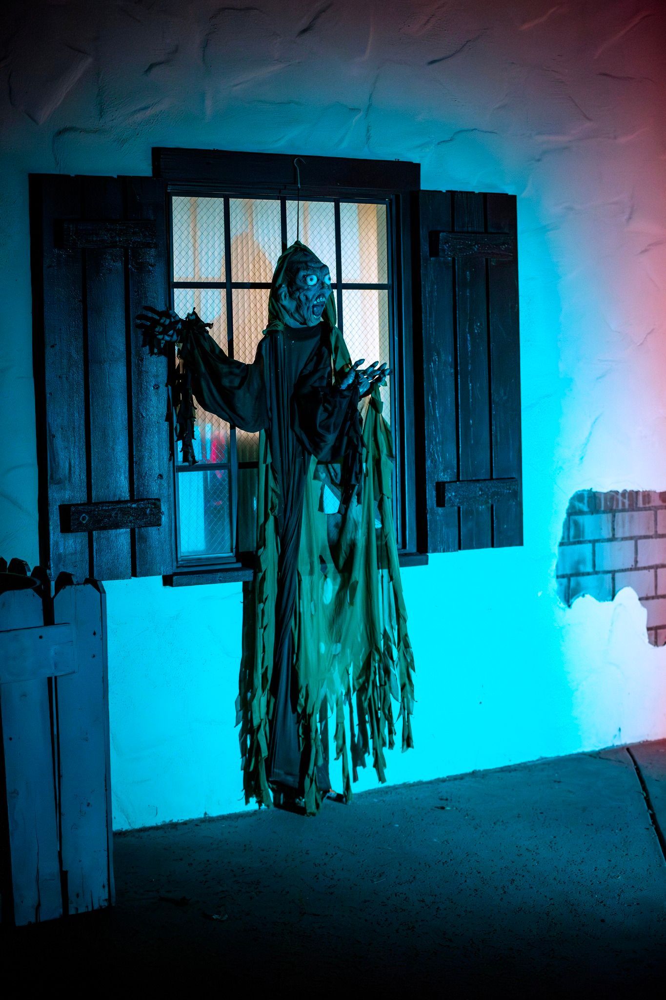 Person in a tattered costume stands in a blue-lit window, holding the shutters open.
