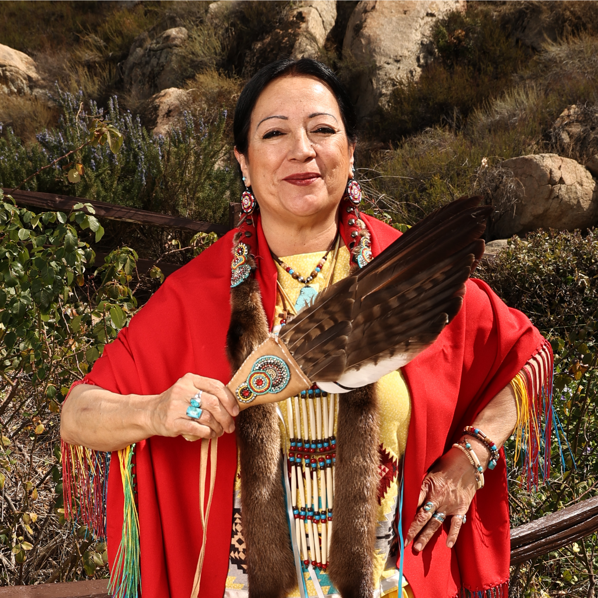 Woman in a red shawl holding a decorated feather fan outdoors among rocks and greenery