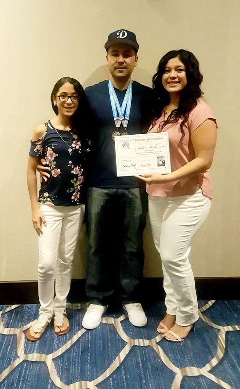 Man in cap stands with two women, holding award. All smiling, posing near a wall, at an event.