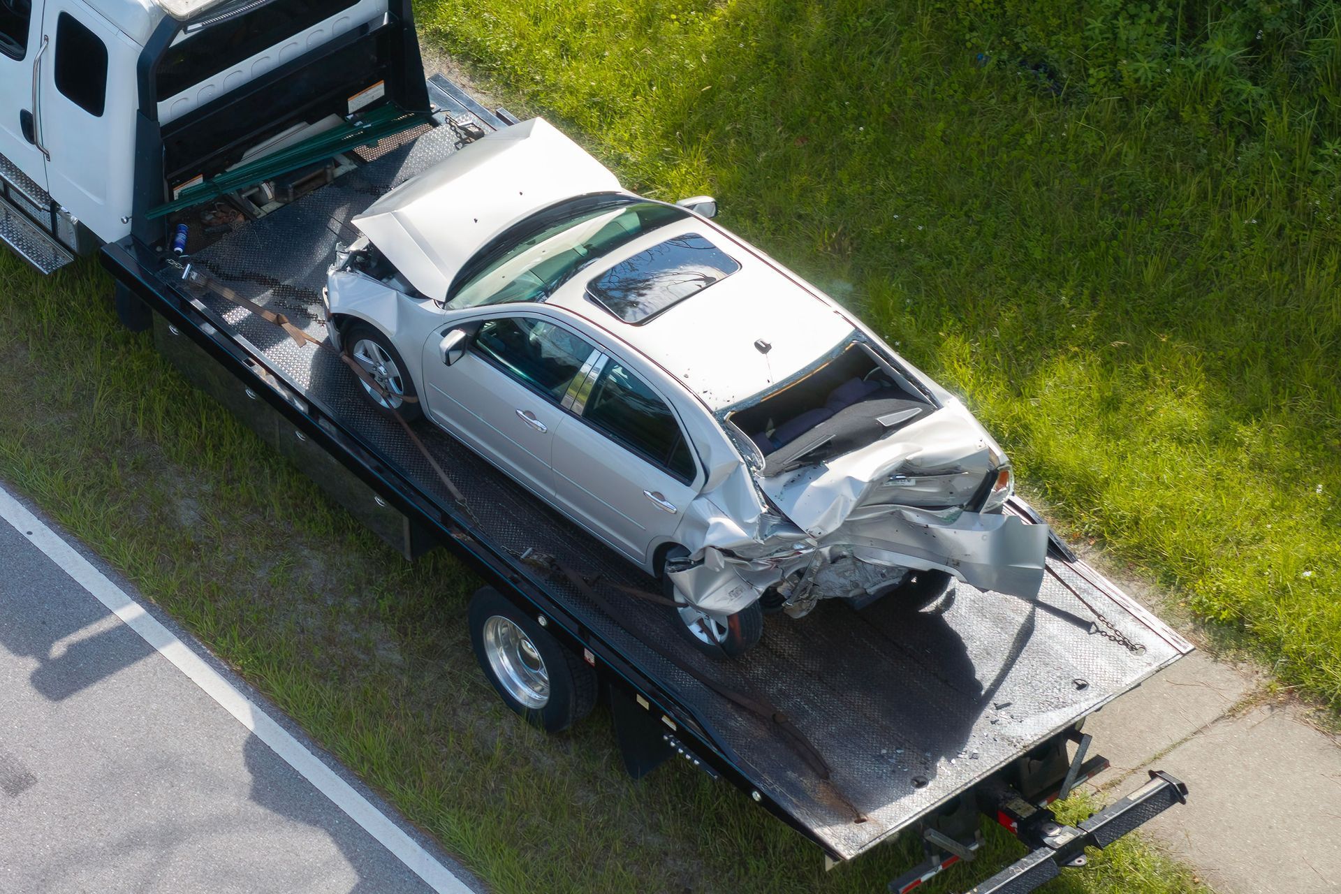 Silver car damaged in a collision, being towed on a flatbed truck next to a grassy roadside.