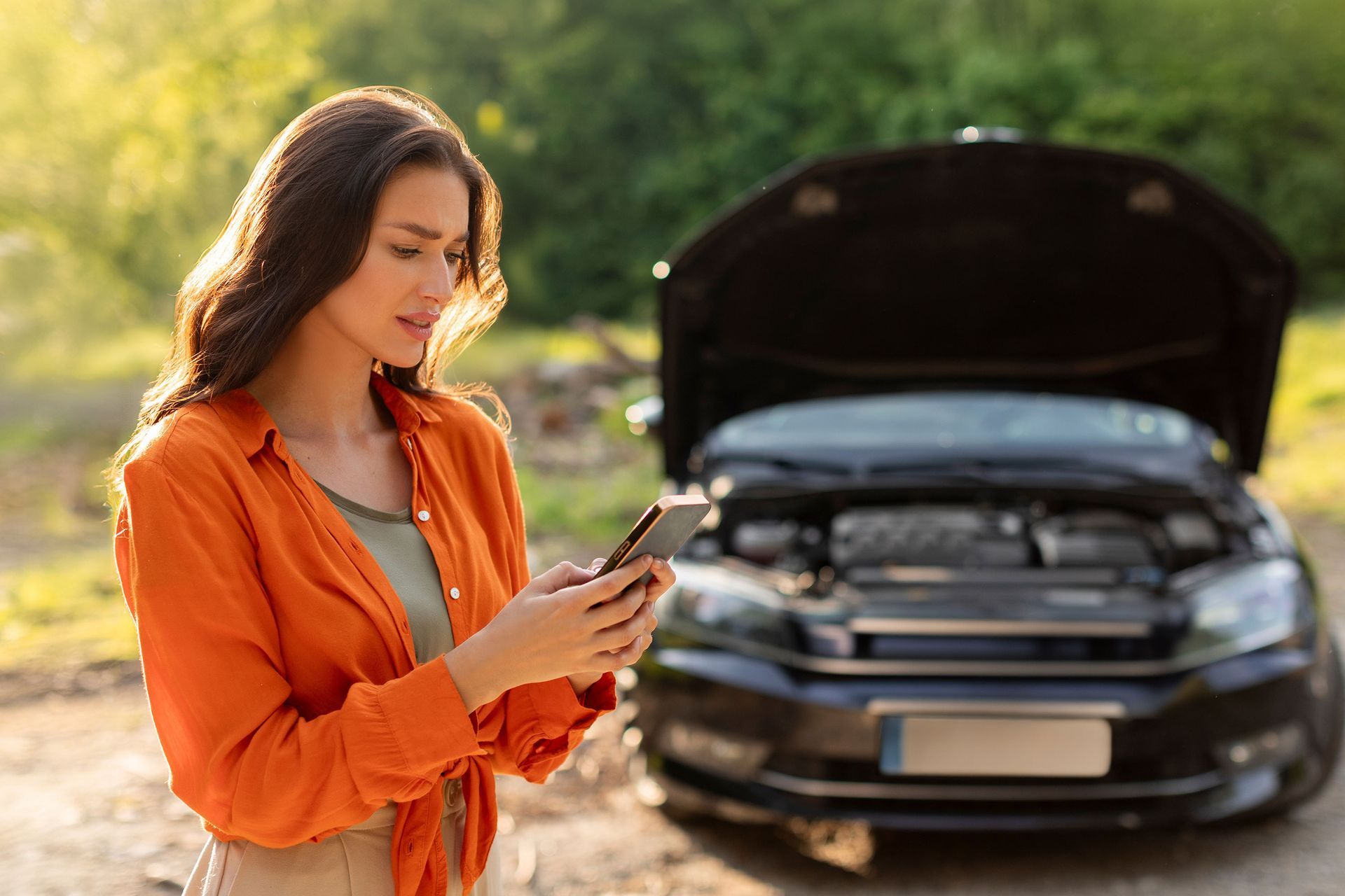 Woman with concerned expression looking at her phone next to a car with an open hood, outdoors.