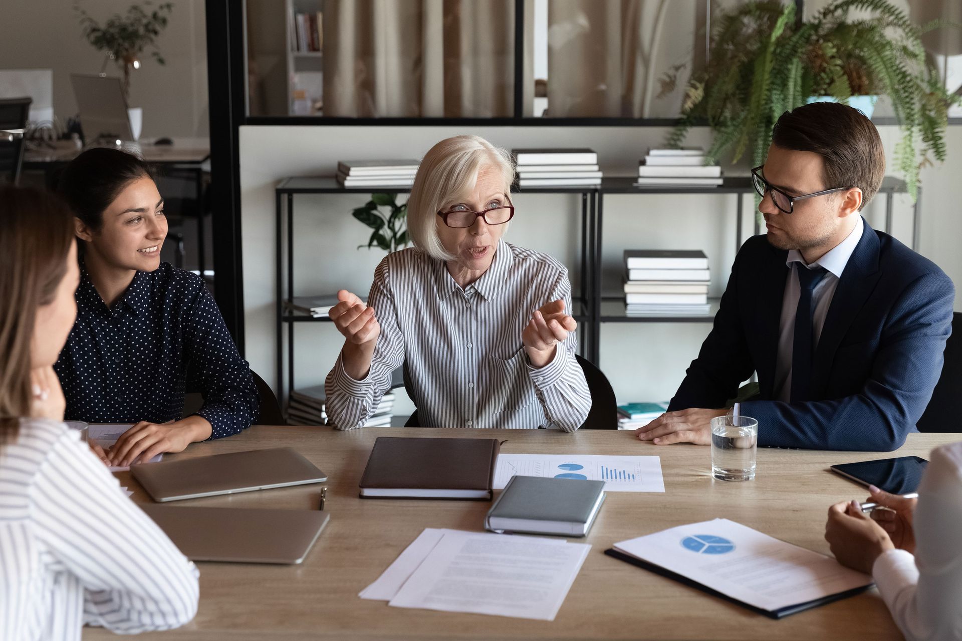 Business meeting with five people around a table, discussing a document. The woman in the center is speaking, and others appear engaged.
