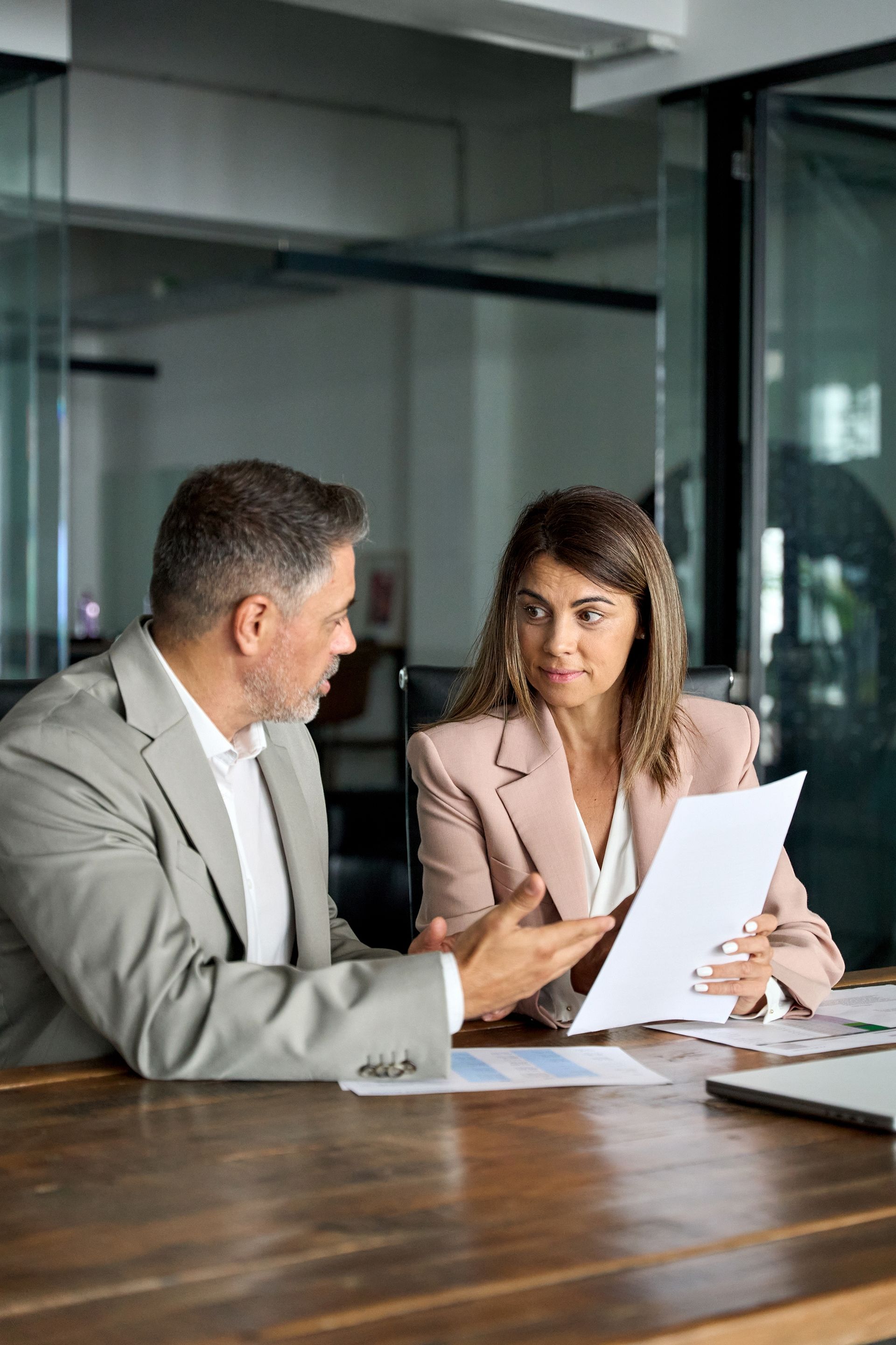 Two businesspeople reviewing documents at a wooden table in an office. Woman points at paper; man gestures with hand.
