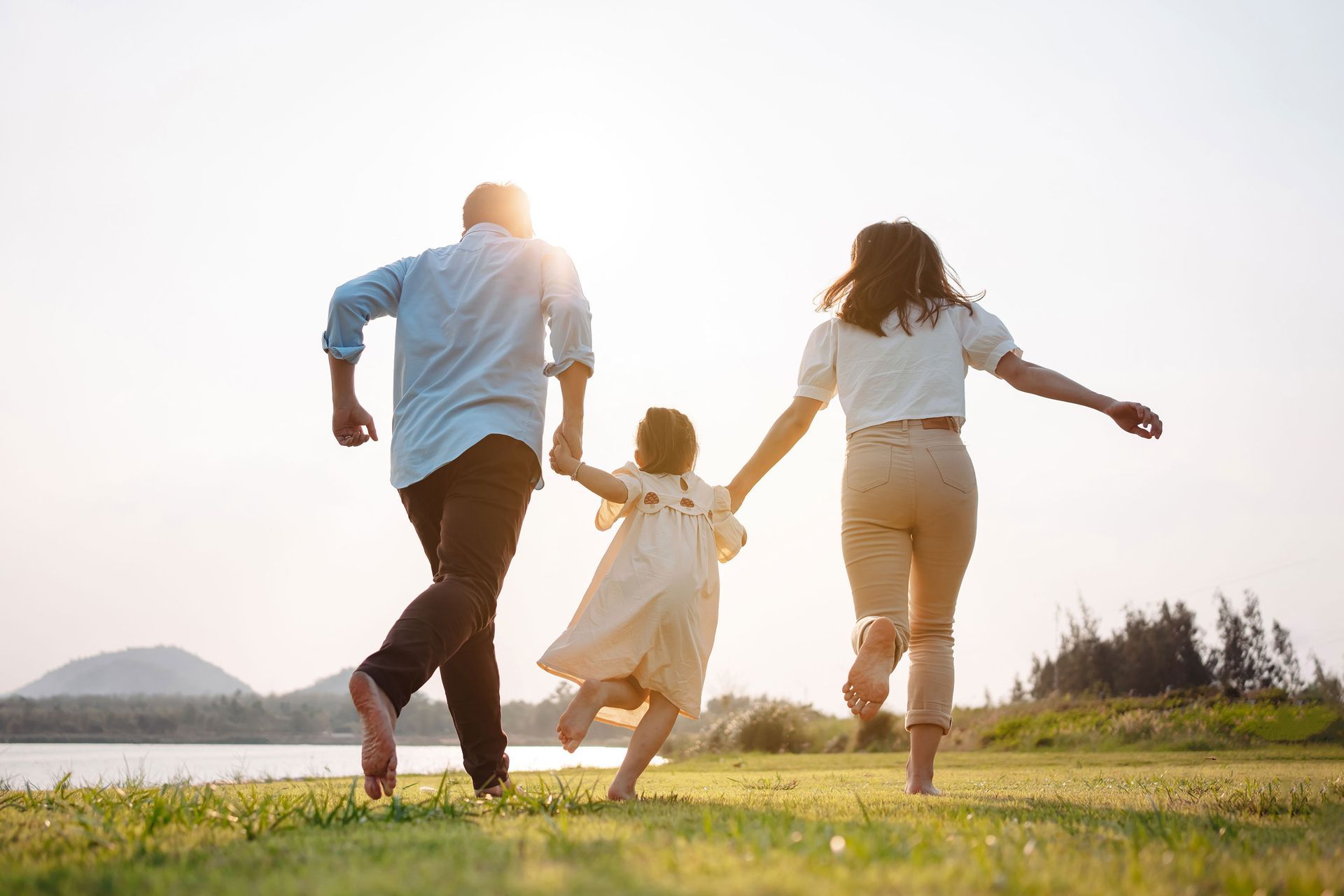 Family running barefoot on a grassy field, holding hands, with a lake and sunlight in the background.