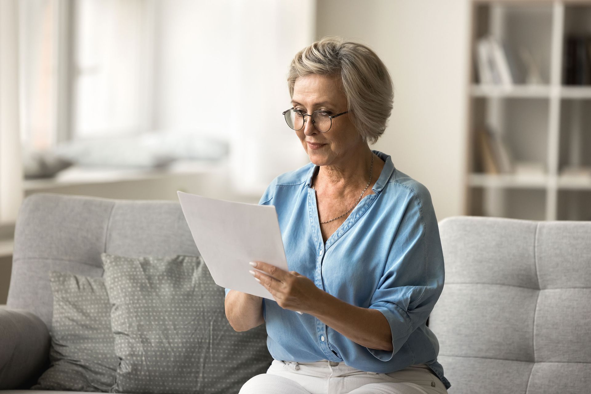 An older woman with glasses sits on a couch, reading a document. She wears a blue shirt and white pants, with a soft expression.