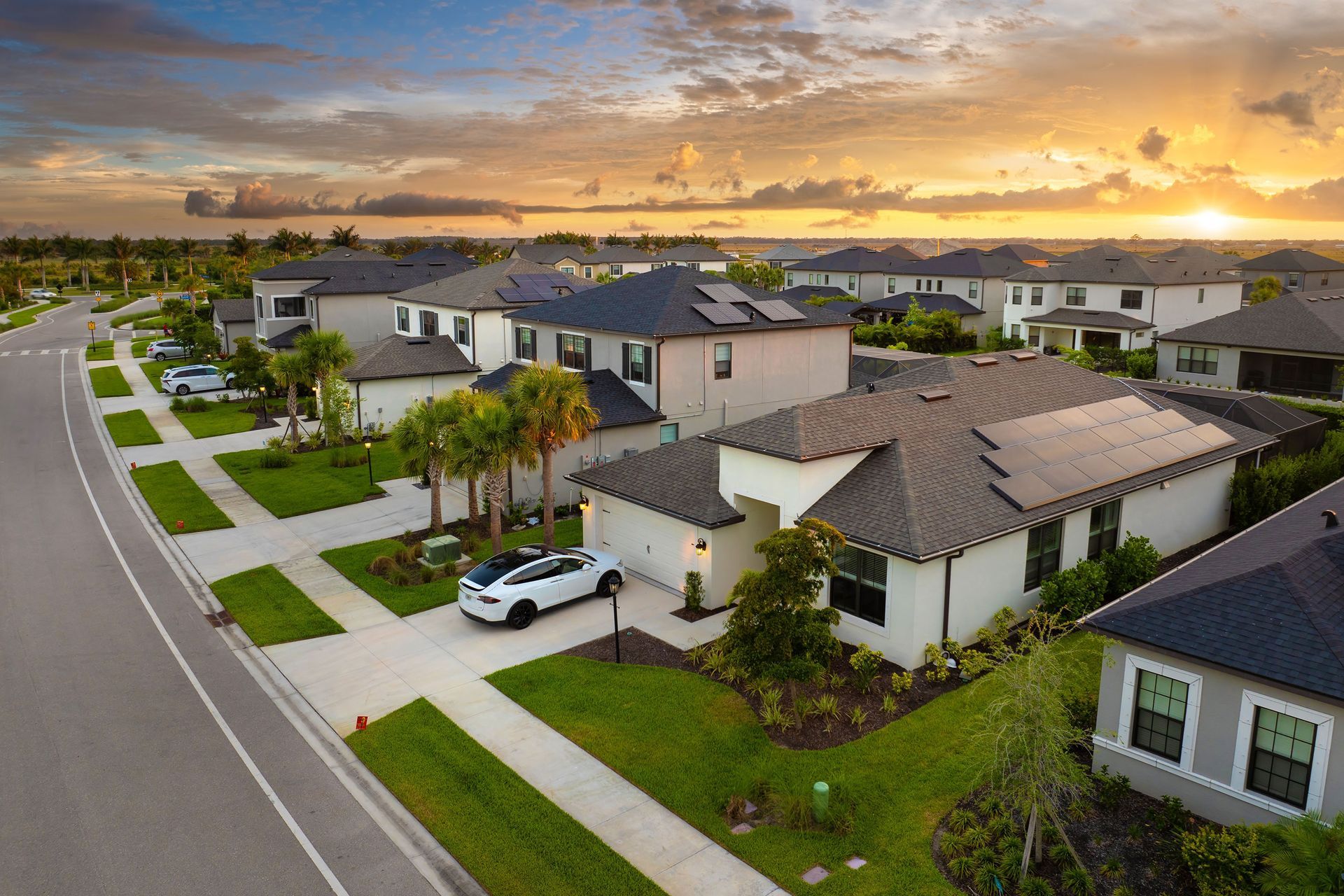 Suburban homes at sunset with a curving road and manicured lawns. A white SUV is parked in front of a house.