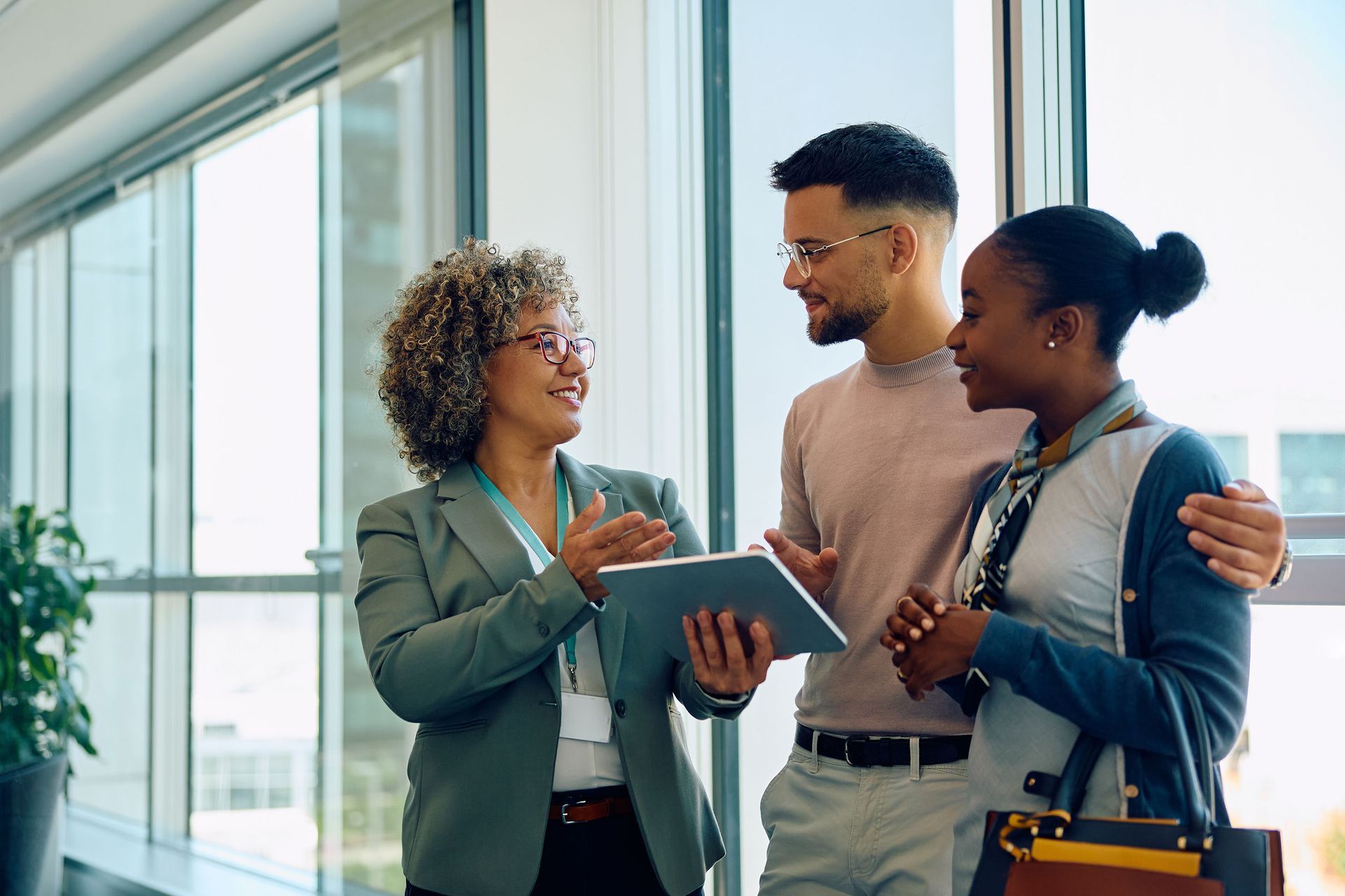 Real estate agent showing a tablet to a smiling couple in a bright modern hallway. The agent gestures with her hands as she speaks.