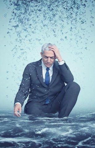 Man in a dark suit kneels in churning water, head in hand, with debris swirling behind him.