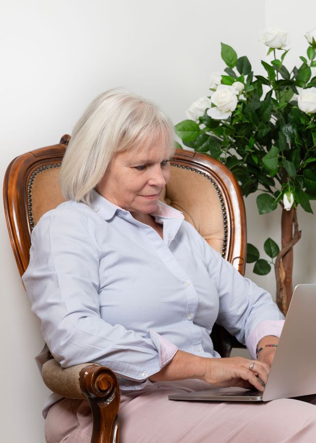 Woman seated in an armchair, typing on a laptop beside a flowering plant.