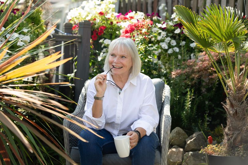 Woman in a white shirt sitting in a flower-filled garden, holding a mug and smiling.