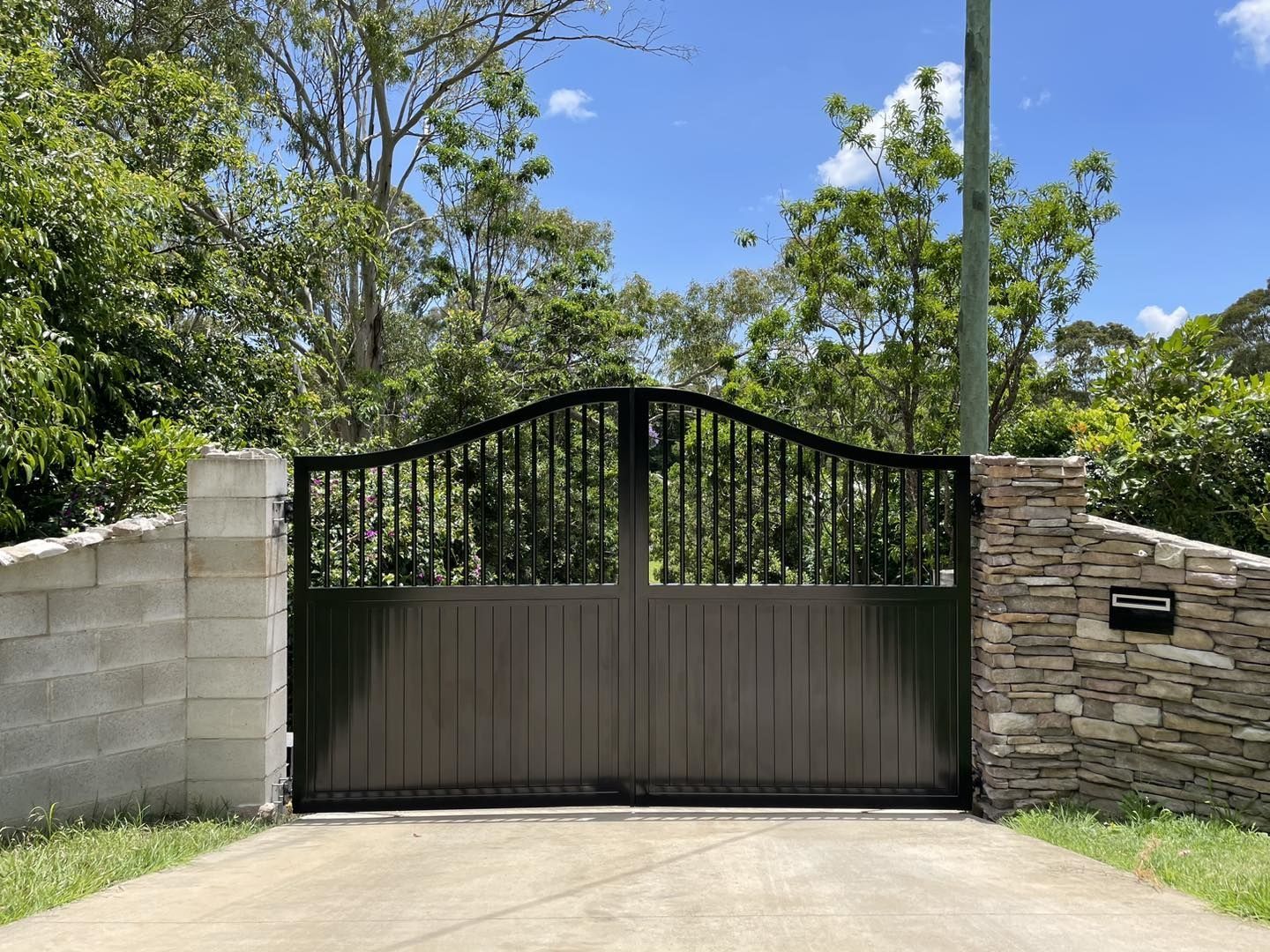 A black gate is sitting in the middle of a driveway next to a stone wall.