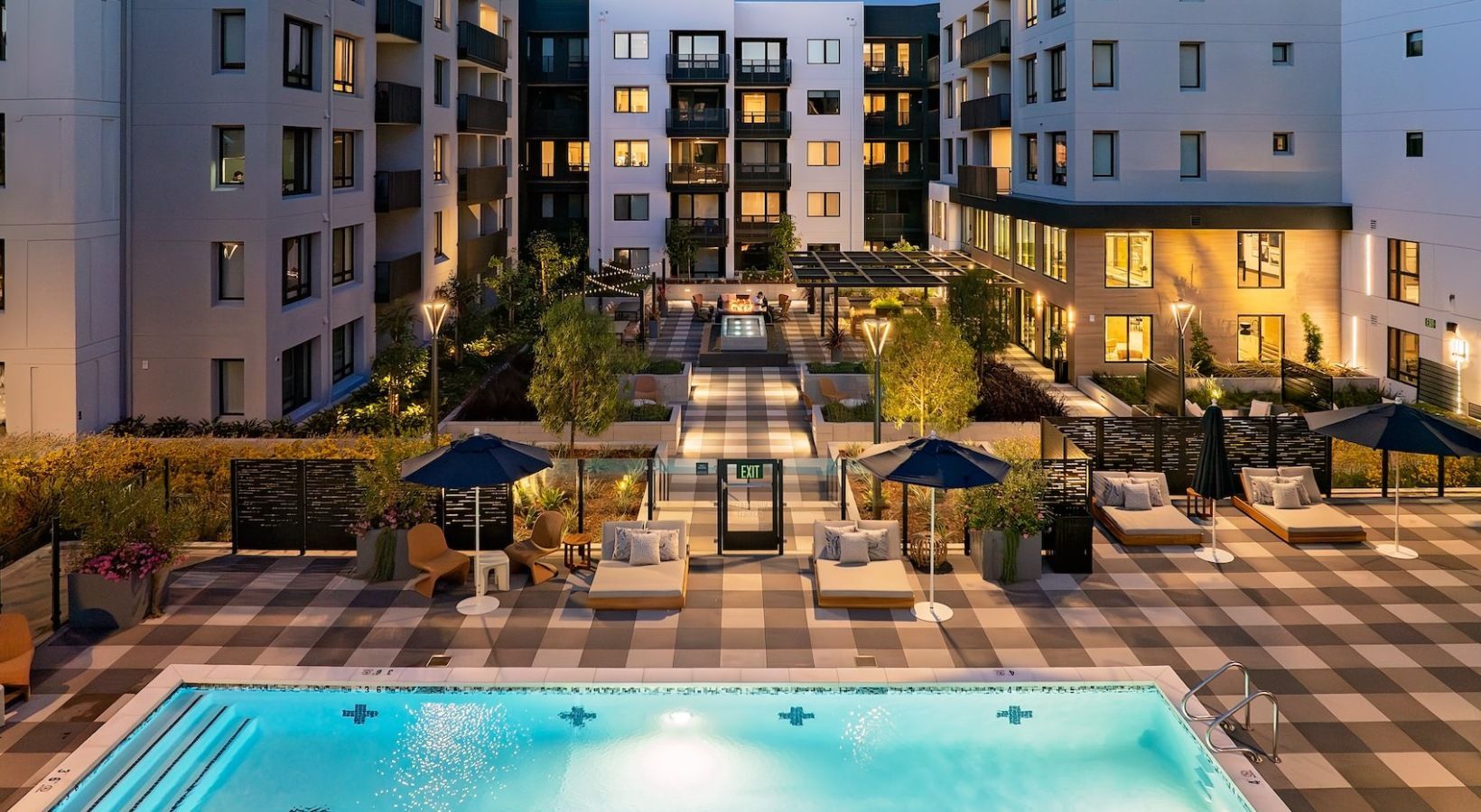 Evening courtyard view of a modern apartment complex with a central pool, lounge chairs, and umbrellas at Sofia, offers Santa Clara apartments for rent in CA.