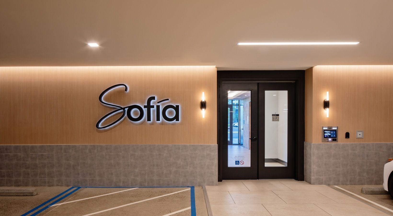 Indoor apartment building lobby with a 'Sofia' sign, double glass doors, and wall sconces.