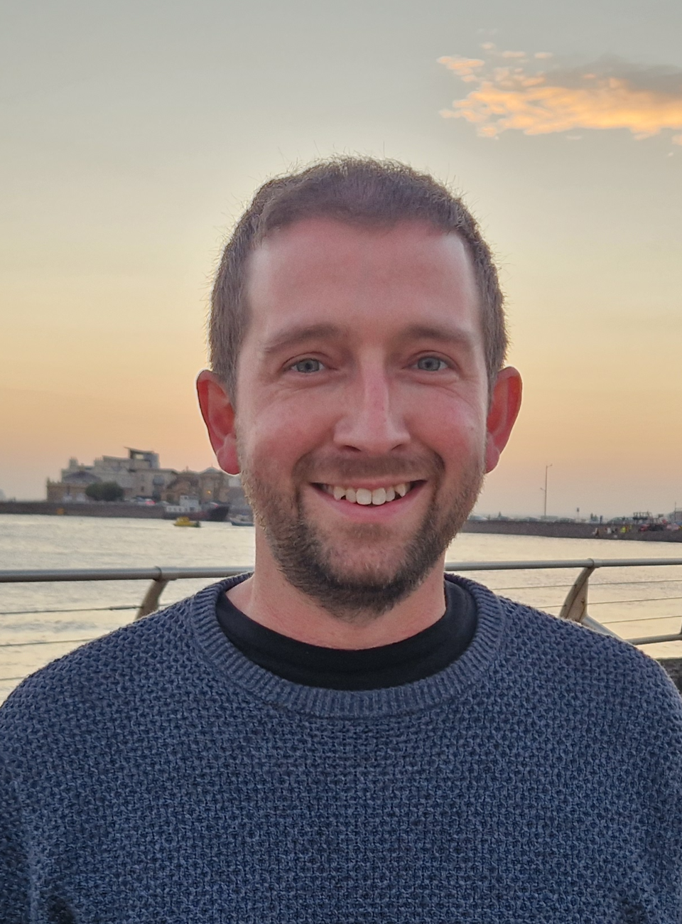 Smiling man in a blue sweater by the water at sunset with a skyline in the background