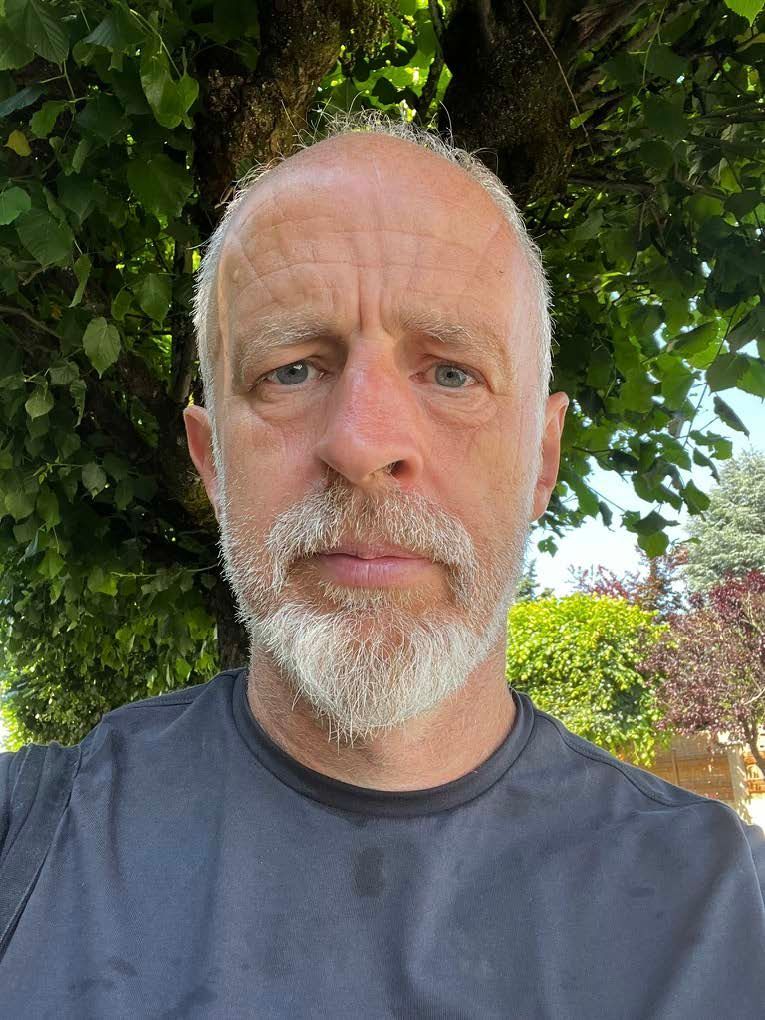 Close-up of a man with a grey beard standing outdoors under leafy trees.