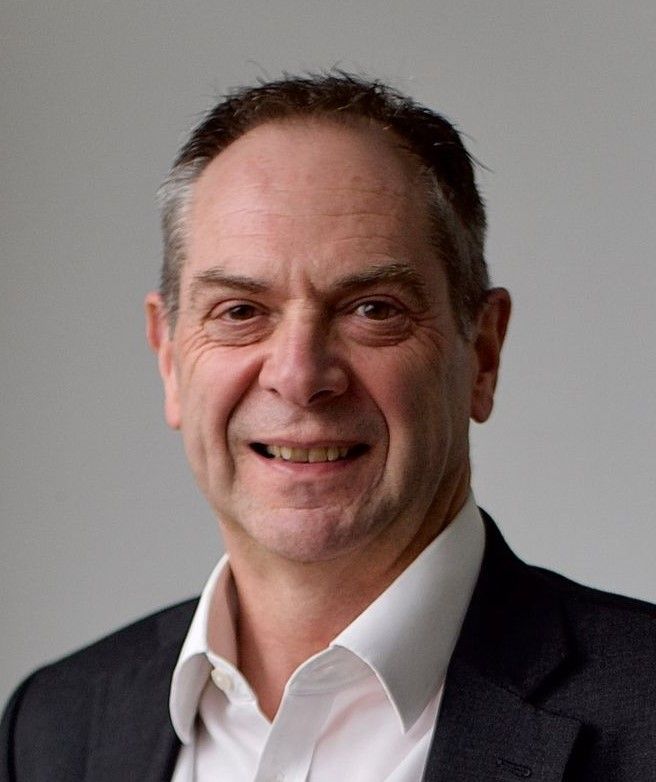 Portrait of a smiling man in a dark suit and white shirt against a plain gray background