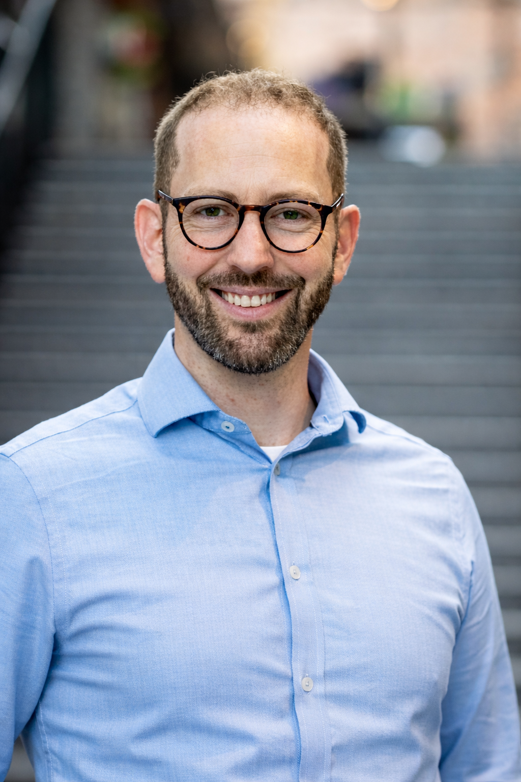 Man with glasses and beard smiles, wearing a light blue button-down shirt, stairs in background.