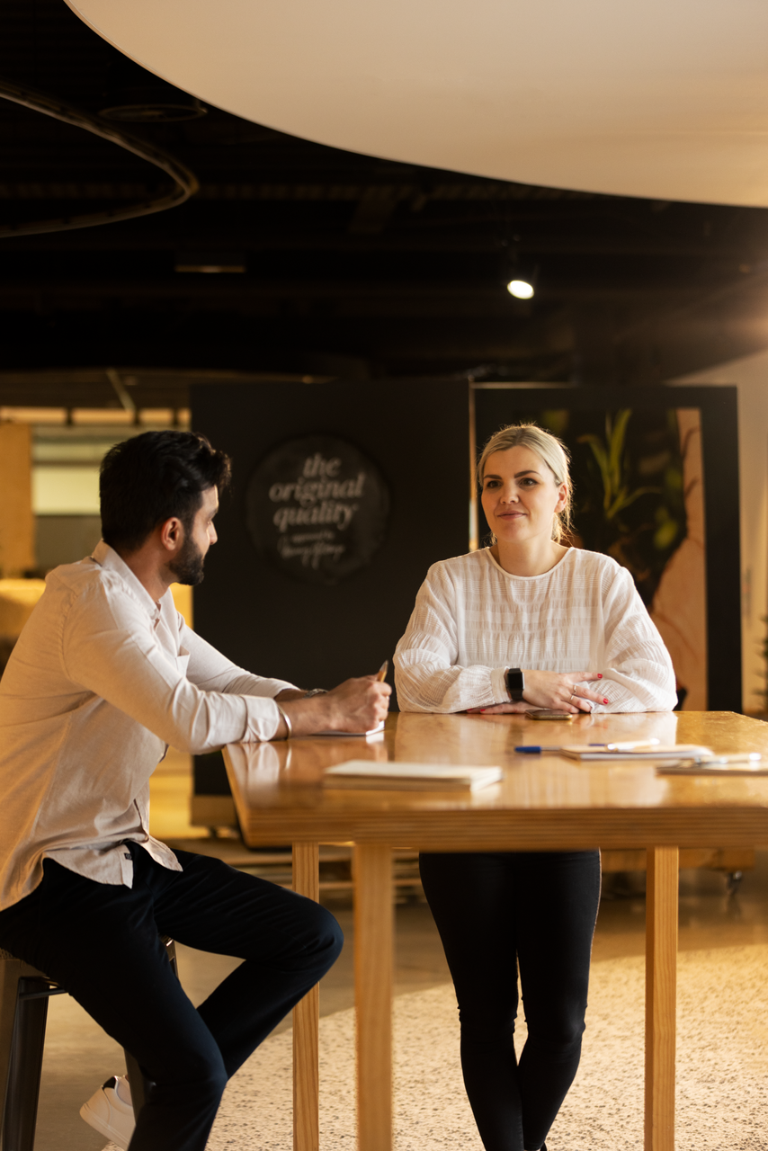 Two people seated at a wooden table in a dim café, talking with drinks in front of them.