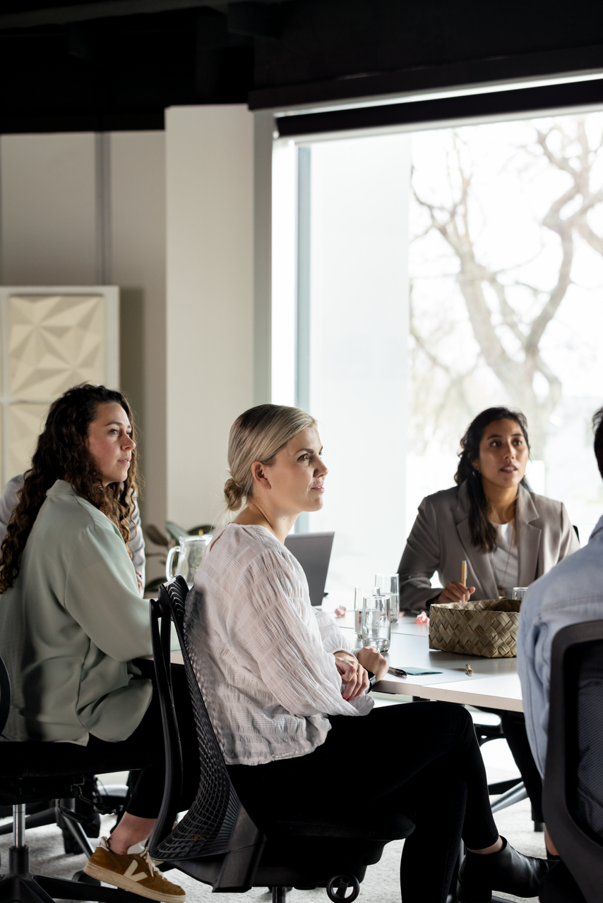 People seated around a table in a bright meeting room, listening and talking during a discussion.