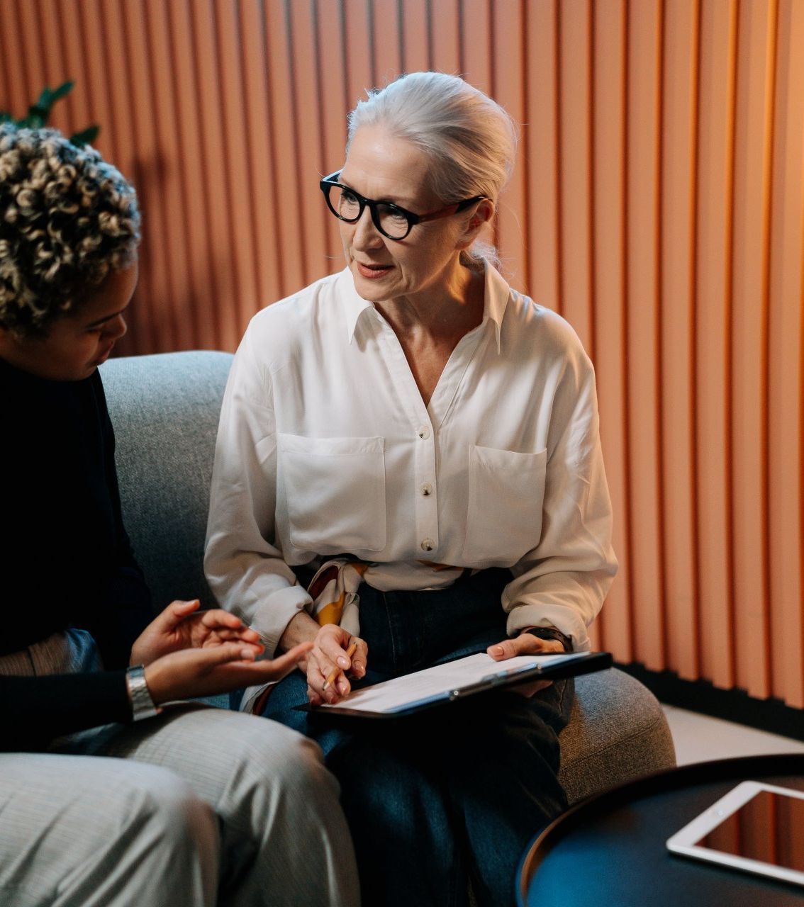 Two people in discussion on a sofa, one holding a notebook in a warm-lit office.
