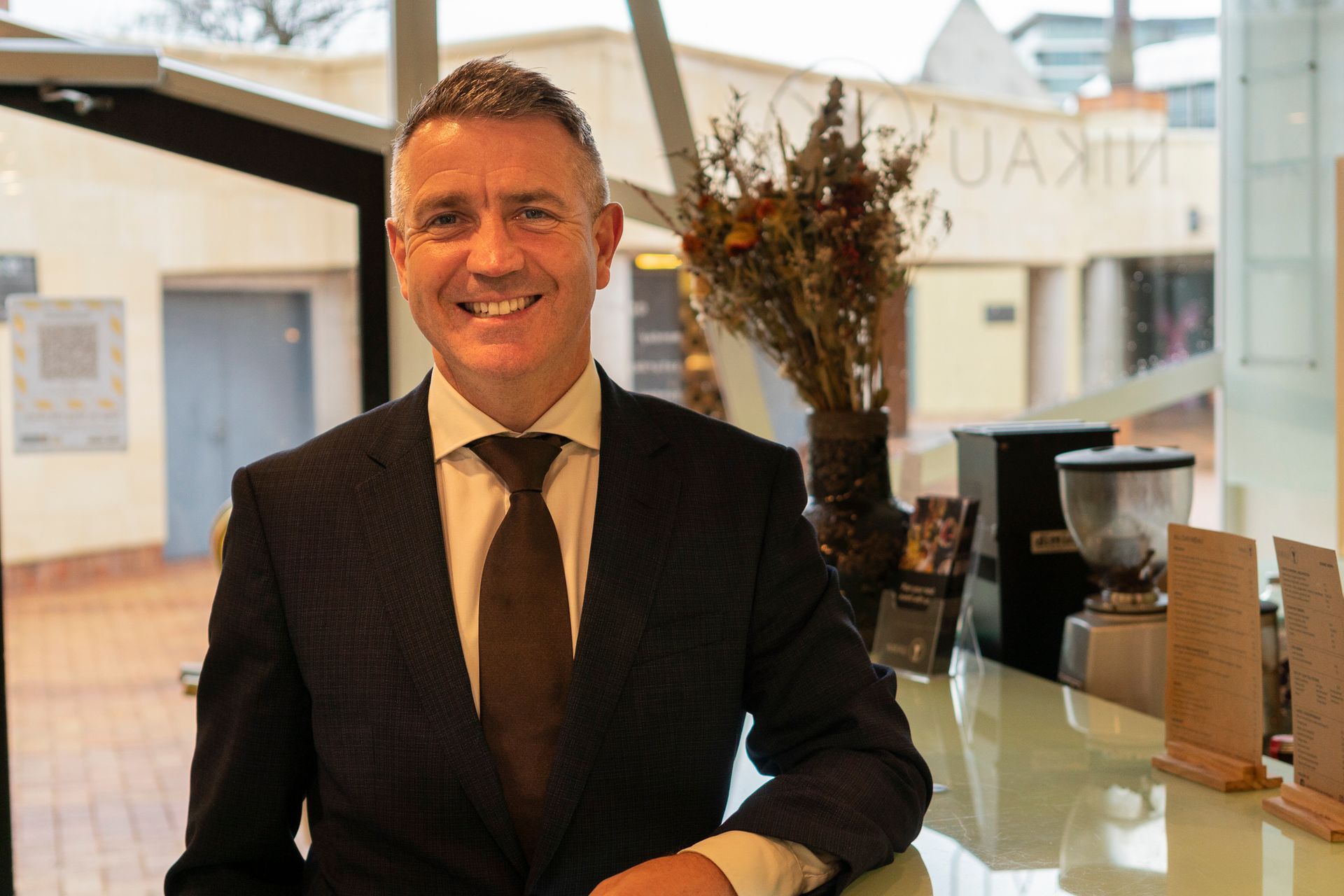 Smiling man in a dark suit standing indoors by a café counter and coffee machine
