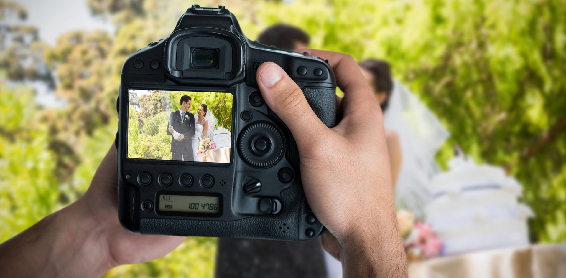Manos sosteniendo una cámara, mostrando una foto de boda en su pantalla, con una pareja borrosa en el fondo.