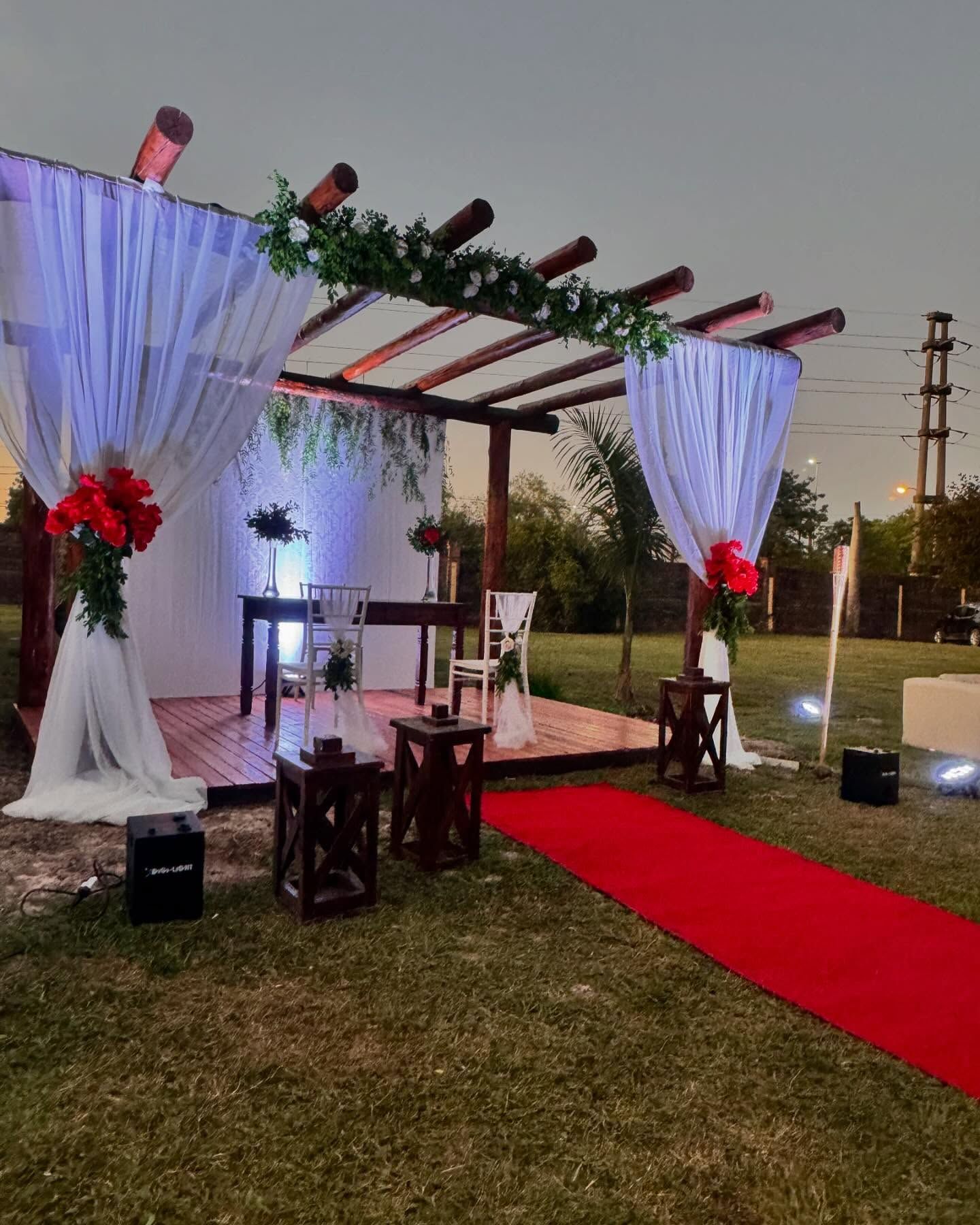 Montaje de ceremonia de boda: pérgola de madera con pasillo de alfombra roja y decoraciones florales; velada al aire libre.
