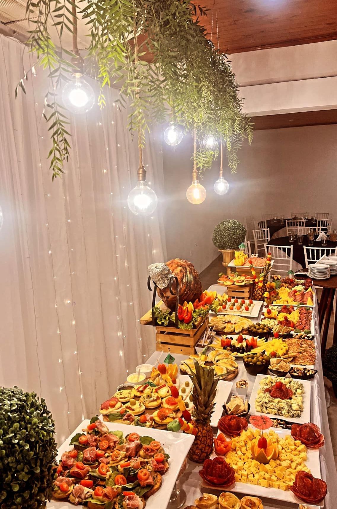 Mesa de buffet cargada de comida bajo luces colgantes y vegetación.