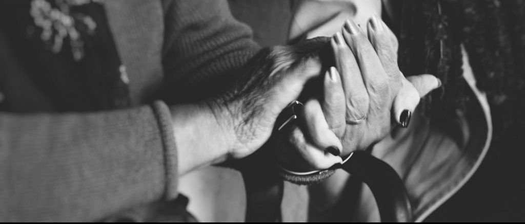 duas mulheres com as mãos dadas. o foco da imagem são as mãos das mulheres, a mulher mais madura usa esmaltes mais claros e da mulher mais nova usa esmaltes escuros