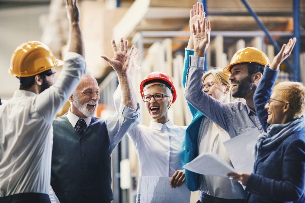 equipe de trabalho comemora conquista. todos estão com suas mãos para cima como se estivessem se preparando para um cumprimento. todos vestem capacete de segurança comum em fábricas
