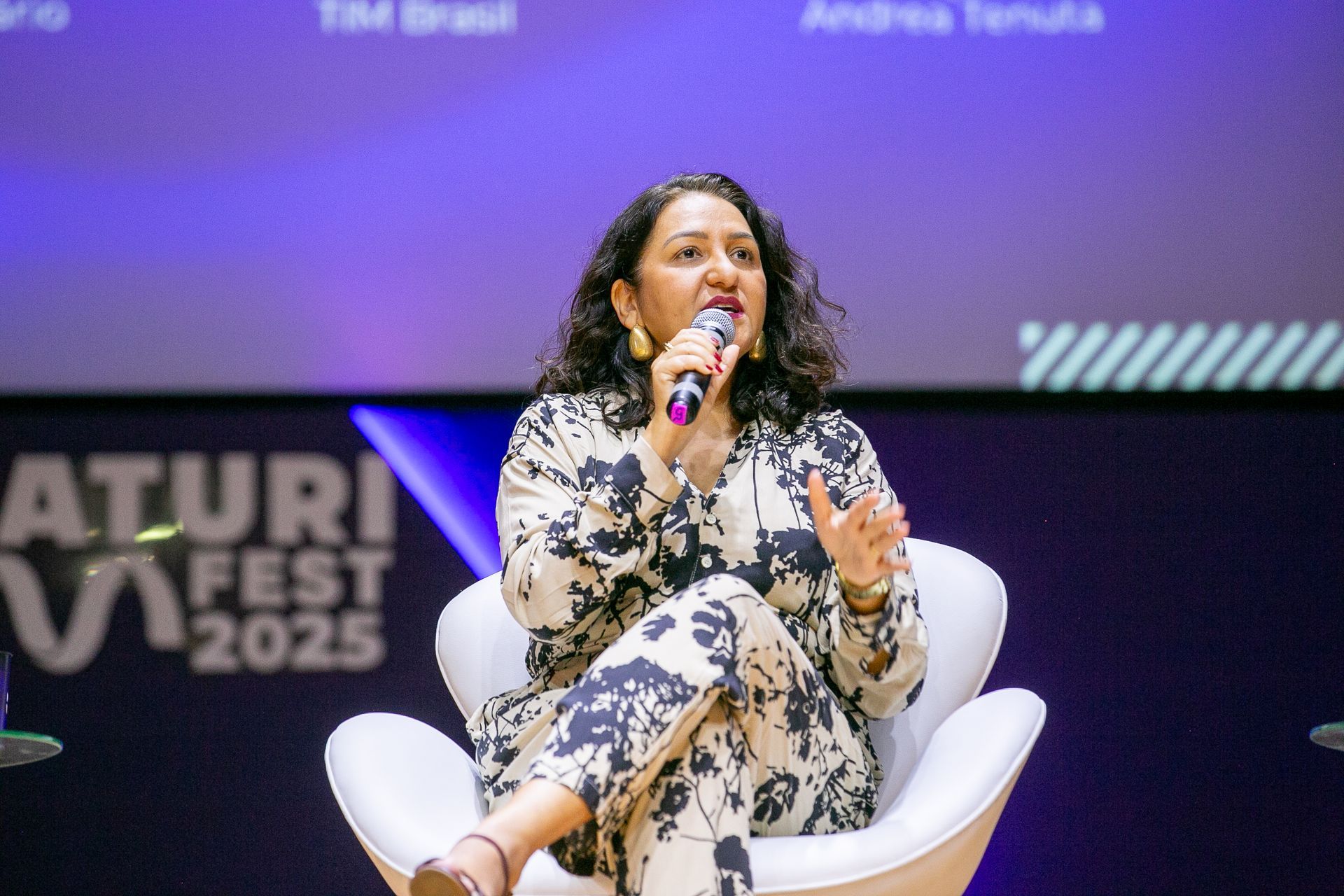 Woman in patterned suit speaking into a microphone at a conference, sitting on a white chair.