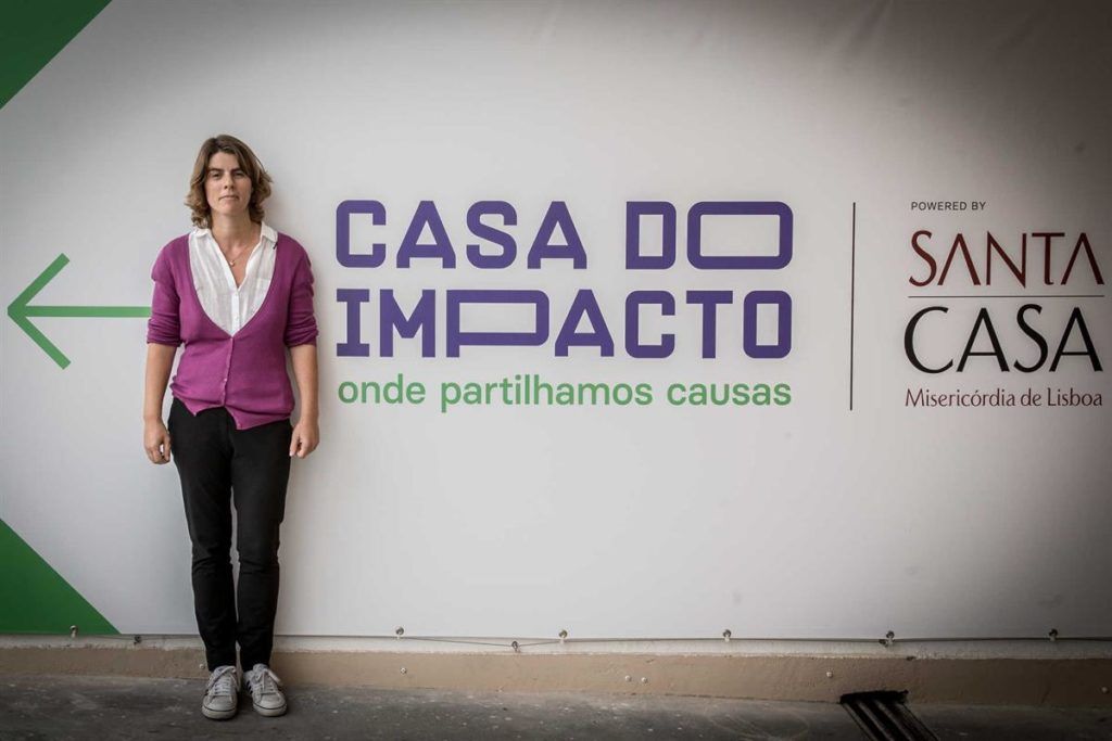mulher posa para foto em frente ao banner da Casa do Impacto. ela veste cardigã roxo, camisa branca e calças pretas. seus cabelos são loiros e curtos e ela não sorri.
